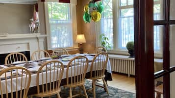 A dining area with a wooden table set for four, surrounded by wooden chairs. There is a chandelier above the table, and a window with curtains to the right.