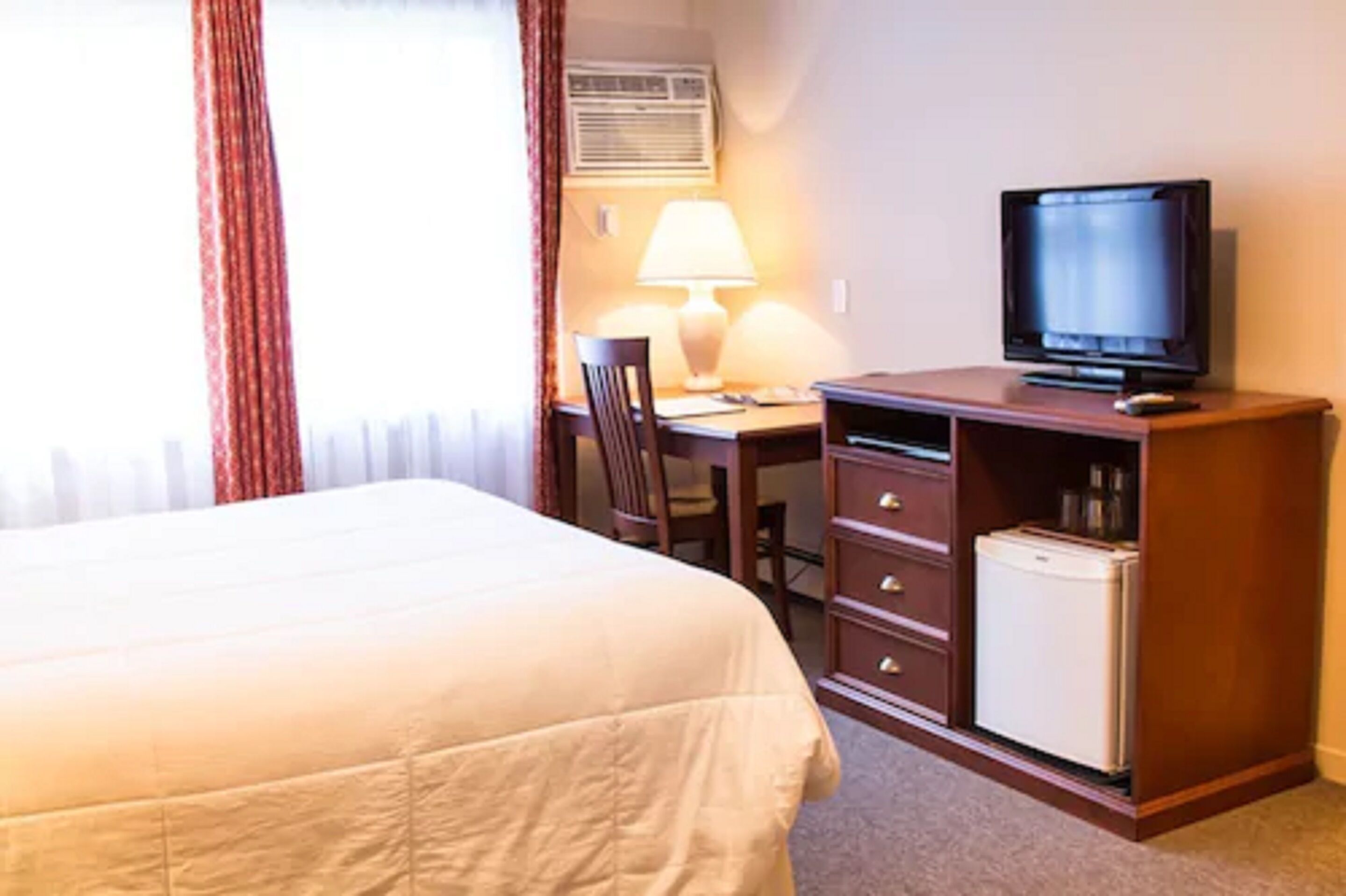 Et hotelværelse med en seng, et skrivebord med lampe, et fjernsyn og et minikøleskab.