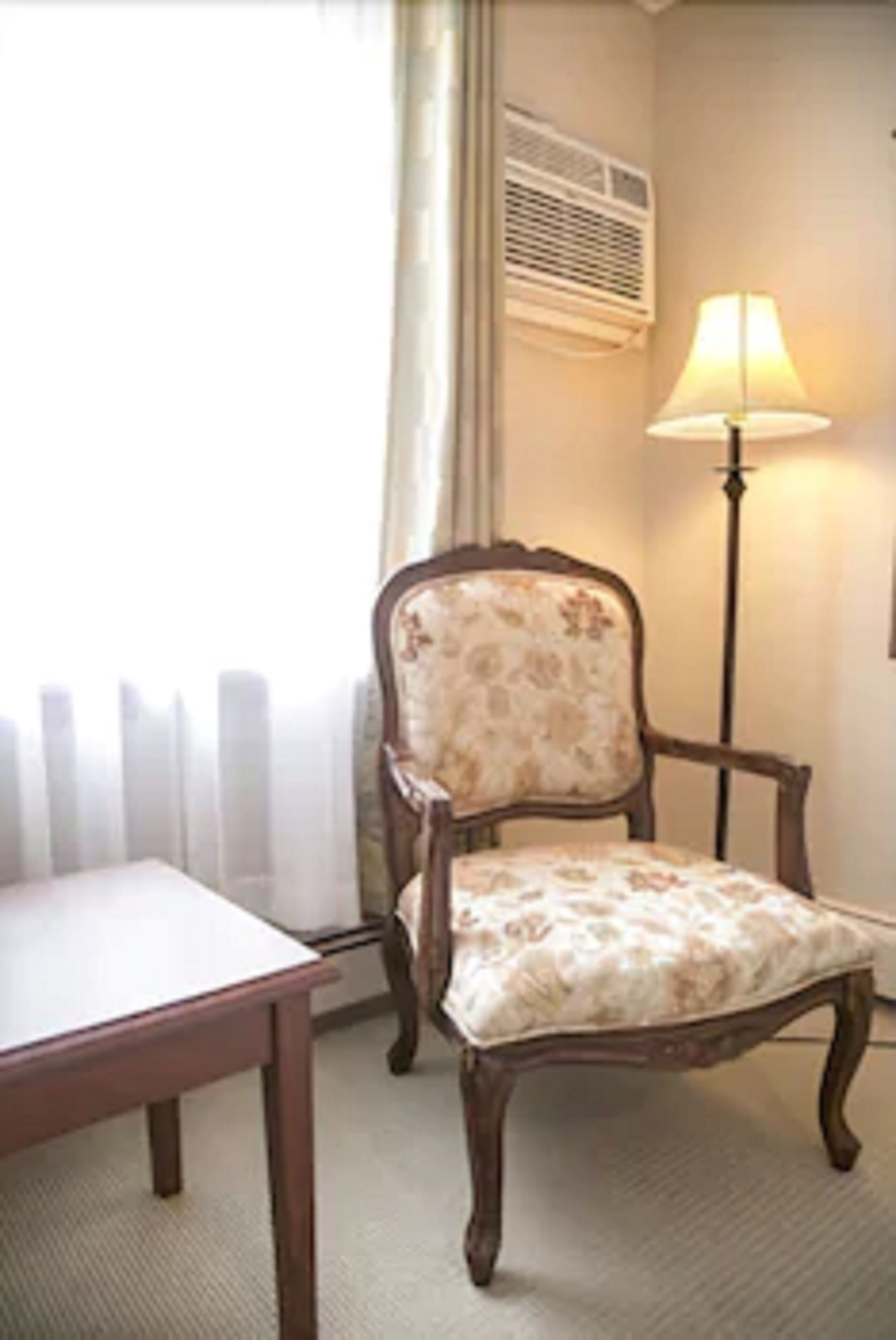 Ein Zimmer mit einem Holzhocker, einem Sitzkissen mit Blumenmuster, einem Tisch, einer Stehlampe und einem Heizkörper.