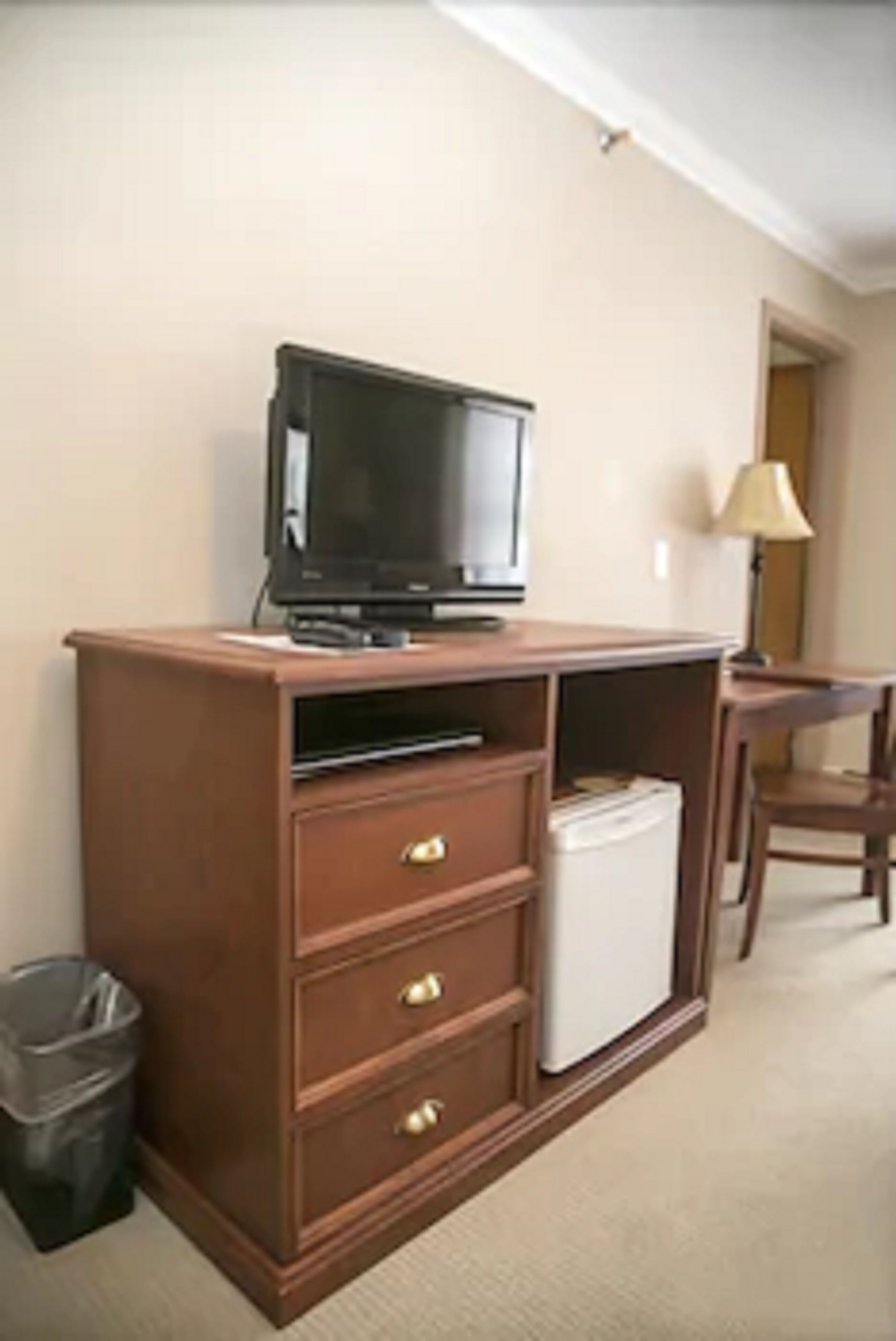 Een kleine kamer met een houten kastje met een televisie, een kleine koelkast en een prullenbak.