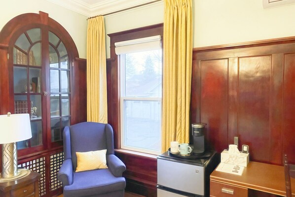 Una habitación con un sillón azul, un armario de madera, una cafetera y una ventana con cortinas.