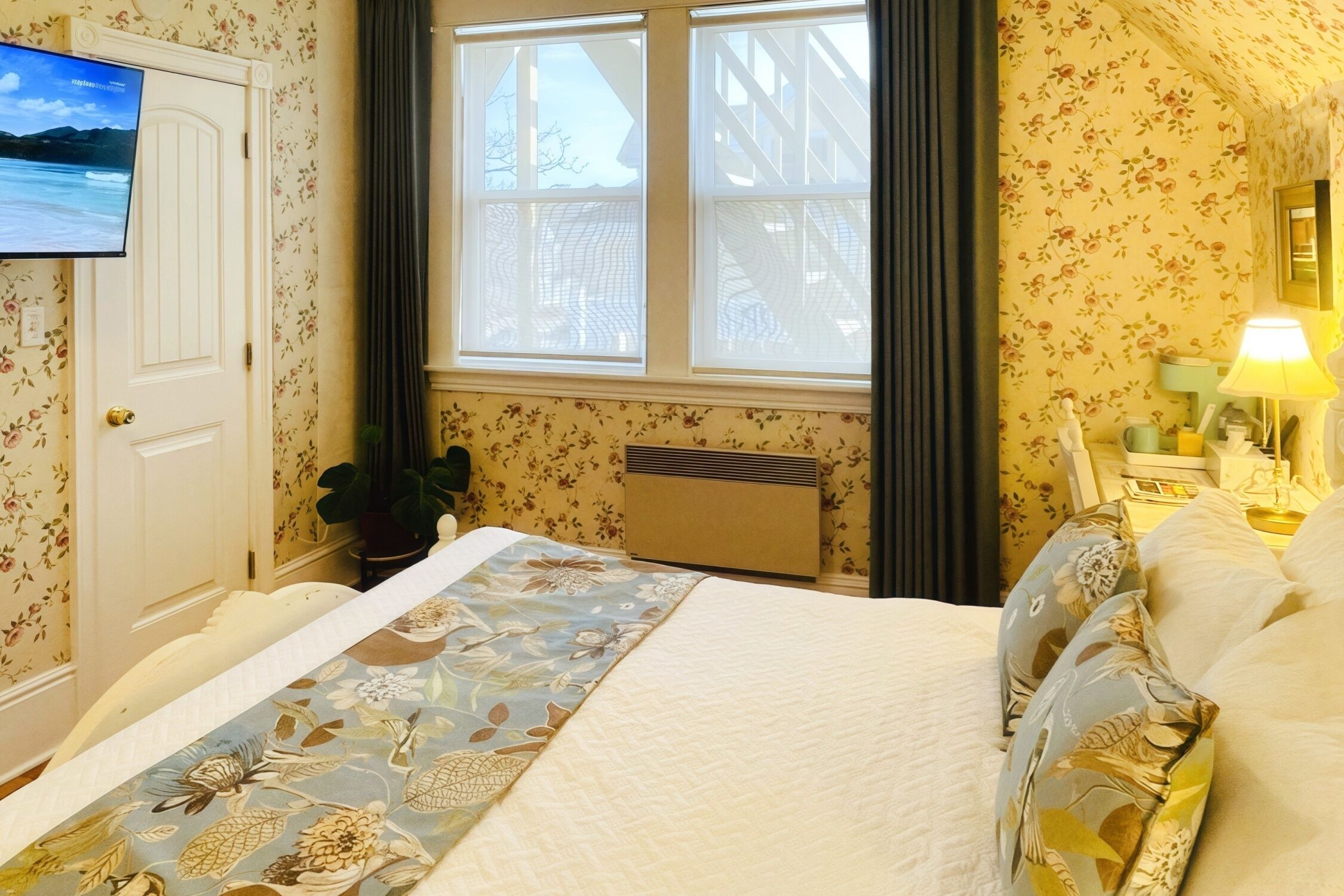 Un dormitorio con cama, televisor en la pared, ventana con cortinas, radiador y mesita de noche con lámpara.