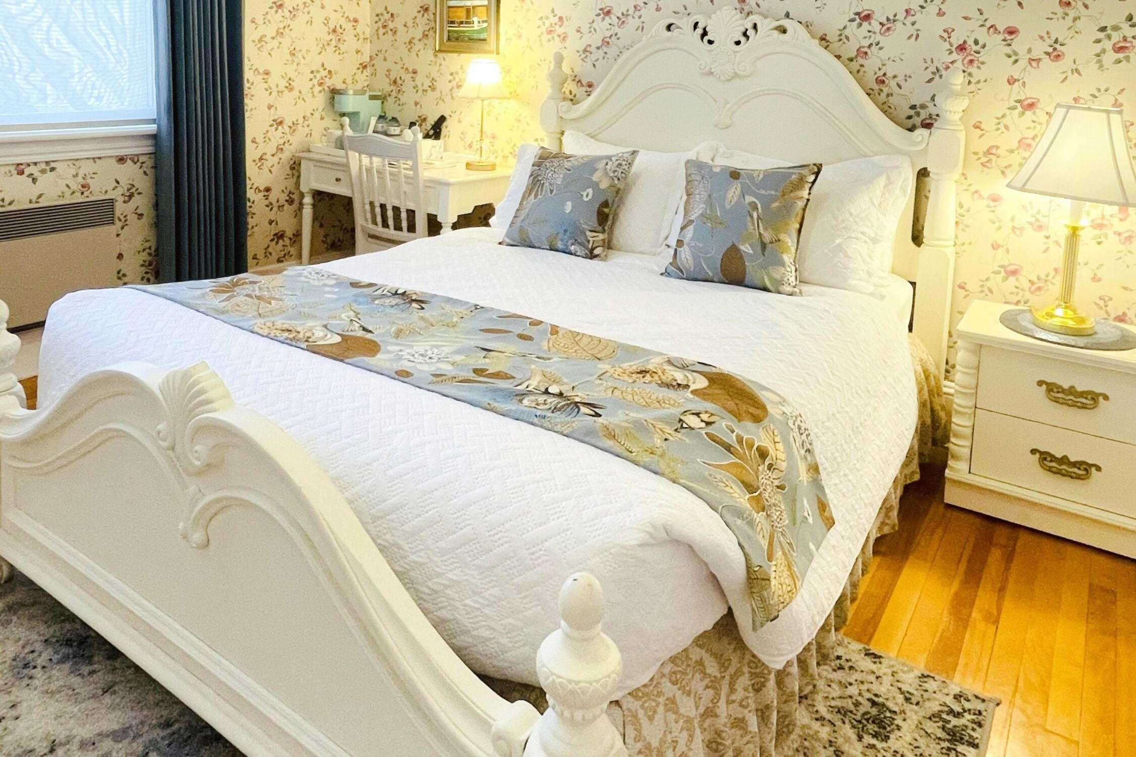 A neatly made bed with a floral bedspread and pillows, a white headboard, a bedside table with a lamp, and a wooden floor.