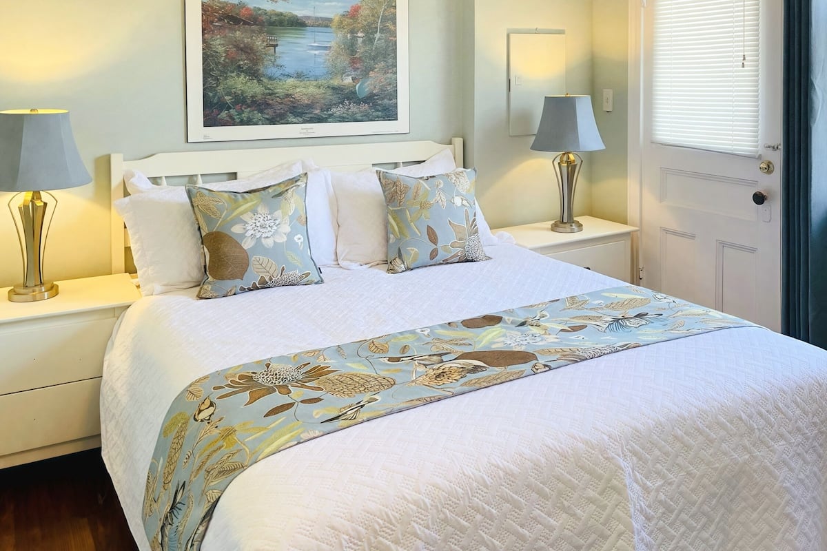 A neatly made bed with a floral bedspread and pillows, a framed landscape painting above the bed, and two bedside lamps on white nightstands.