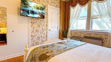 A bedroom with floral wallpaper, a bed with a floral bedspread, a mounted TV, a window with curtains, and a radiator.
