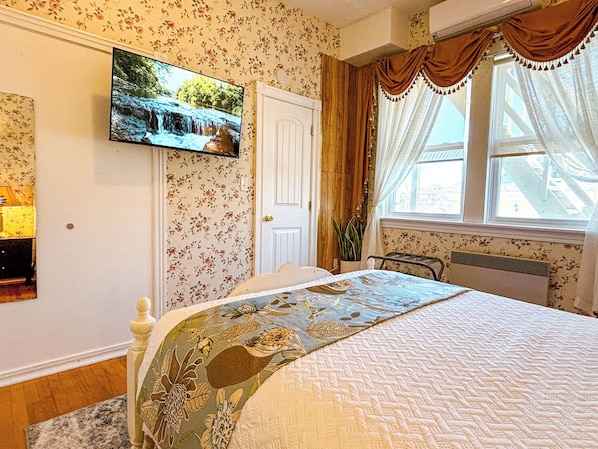 Un dormitorio con papel tapiz floral, una cama con cubrecama floral, un televisor montado, una ventana con cortinas y un radiador.