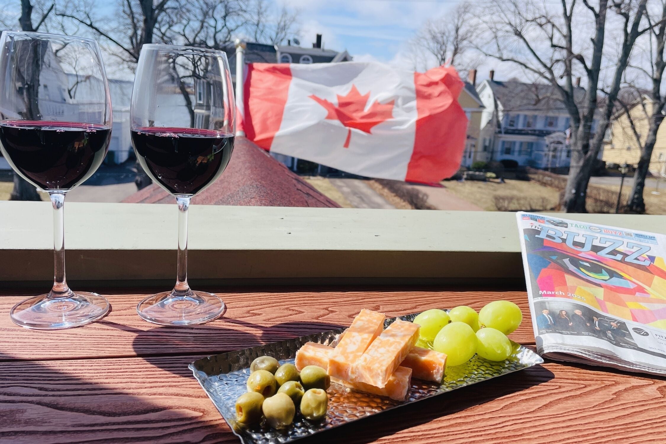 Deux verres de vin rouge, une assiette de fromage et de raisins, et un drapeau canadien en arrière-plan.