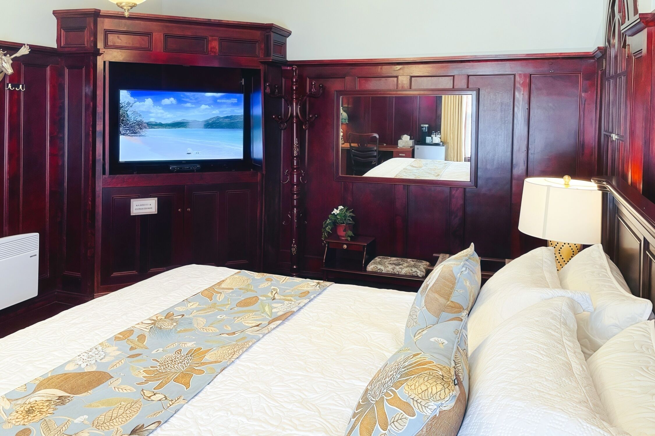 A hotel room with a large bed, a flat-screen TV, a mirror, a bedside table, and a lamp.