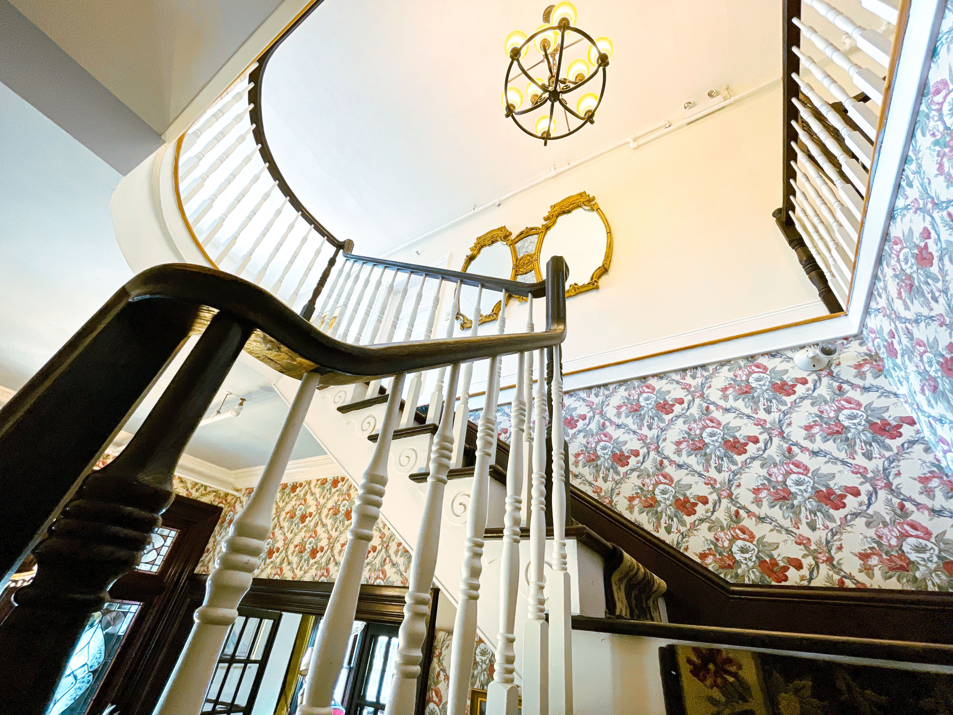 A grand staircase with ornate railings and a chandelier, leading to a room with floral wallpaper and a ceiling with a decorative design.
