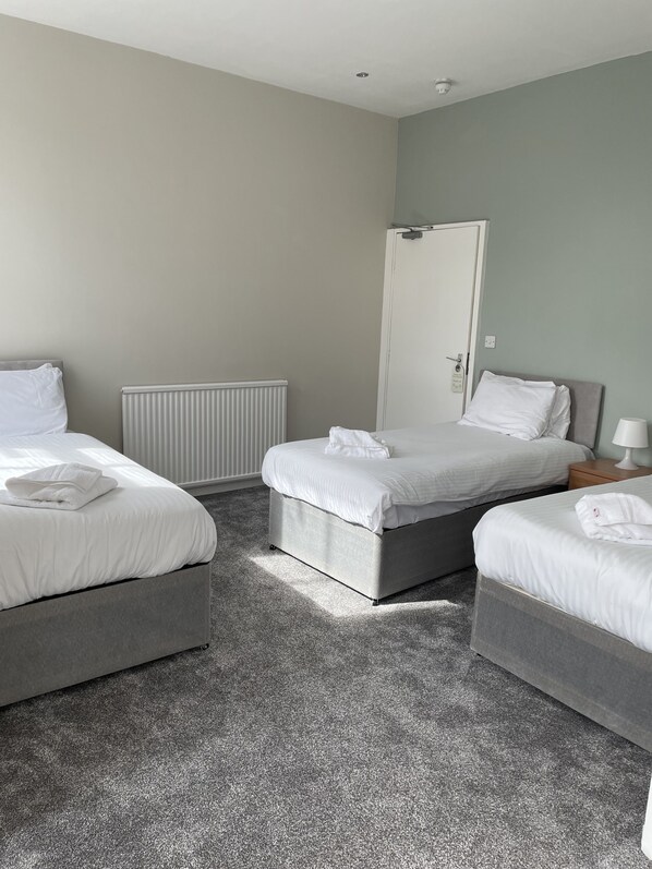 A hotel room with two beds, a radiator, a door, and a nightstand with a lamp. - The Georgian Hollies (Blackpool)