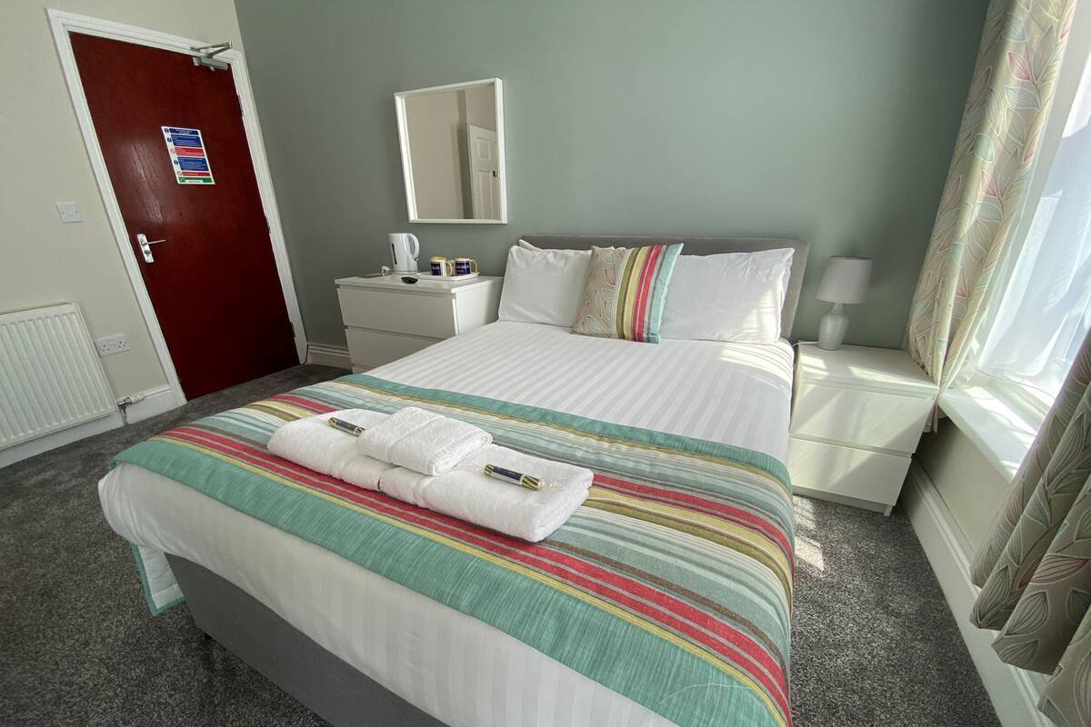 A hotel room with a bed, a nightstand, a mirror, a window with curtains, and a door.