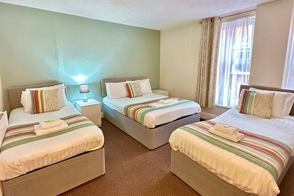 A hotel room with three beds, each with striped bedding and pillows. There is a nightstand with a lamp between the beds. The room has a carpeted floor and a window with curtains.