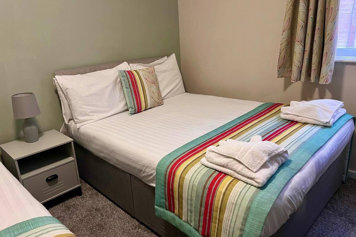 A neatly made bed with a striped blanket, a pillow, and a folded towel on the bed. There is a bedside table with a lamp and a drawer.