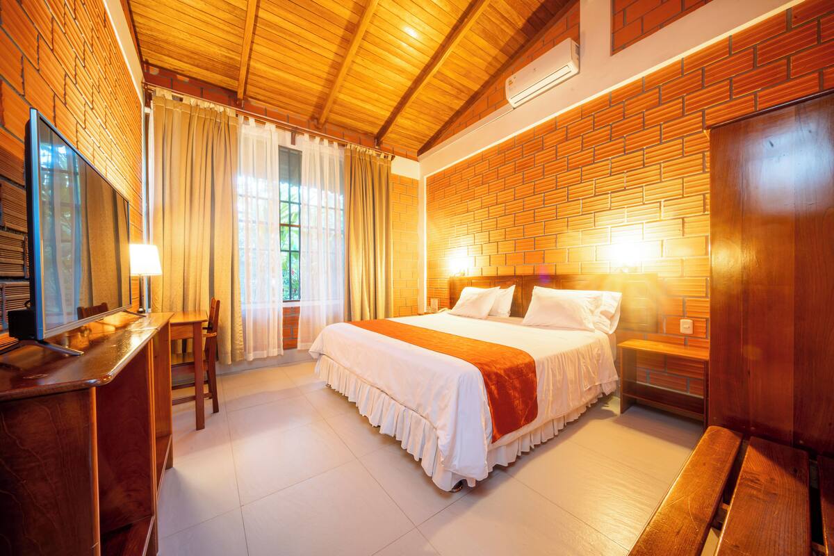 A hotel room with a wooden ceiling, a brick wall, a large bed with white bedding, a desk with a chair, and a window with curtains.