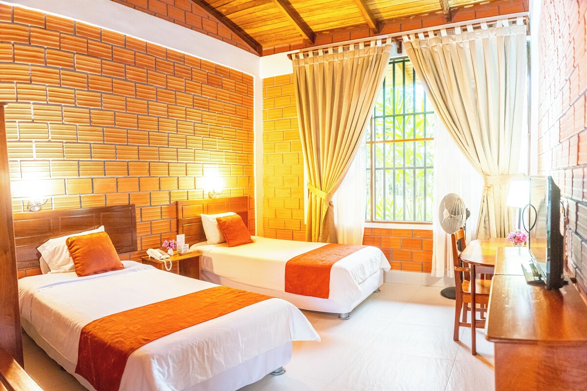 Habitación de hotel con dos camas, techo de madera, paredes de ladrillo, ventana con cortinas, escritorio con lámpara y televisor.