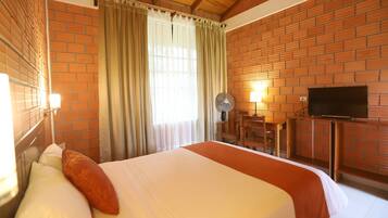 Una habitación de hotel con pared de ladrillo, cama con ropa de cama blanca y naranja, televisor de pantalla plana, escritorio con silla y un ventilador.