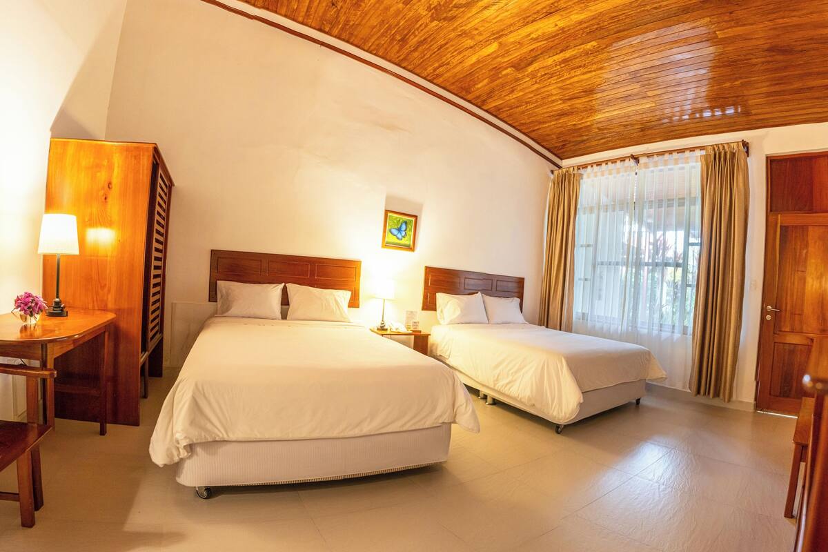 A hotel room with two beds, wooden ceiling, and a window with curtains.