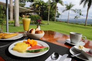 A breakfast setting with plates of fruit, a bowl of bread, a cup of coffee, and a view of a lush landscape. -  Altavista Casahotel - Asociado Casa Andina (Moyobamba)