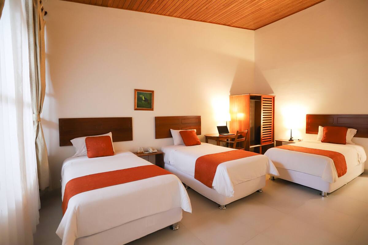 A hotel room with three beds, each with white bedding and orange accents, a wooden ceiling, and a small desk with a lamp and a laptop.