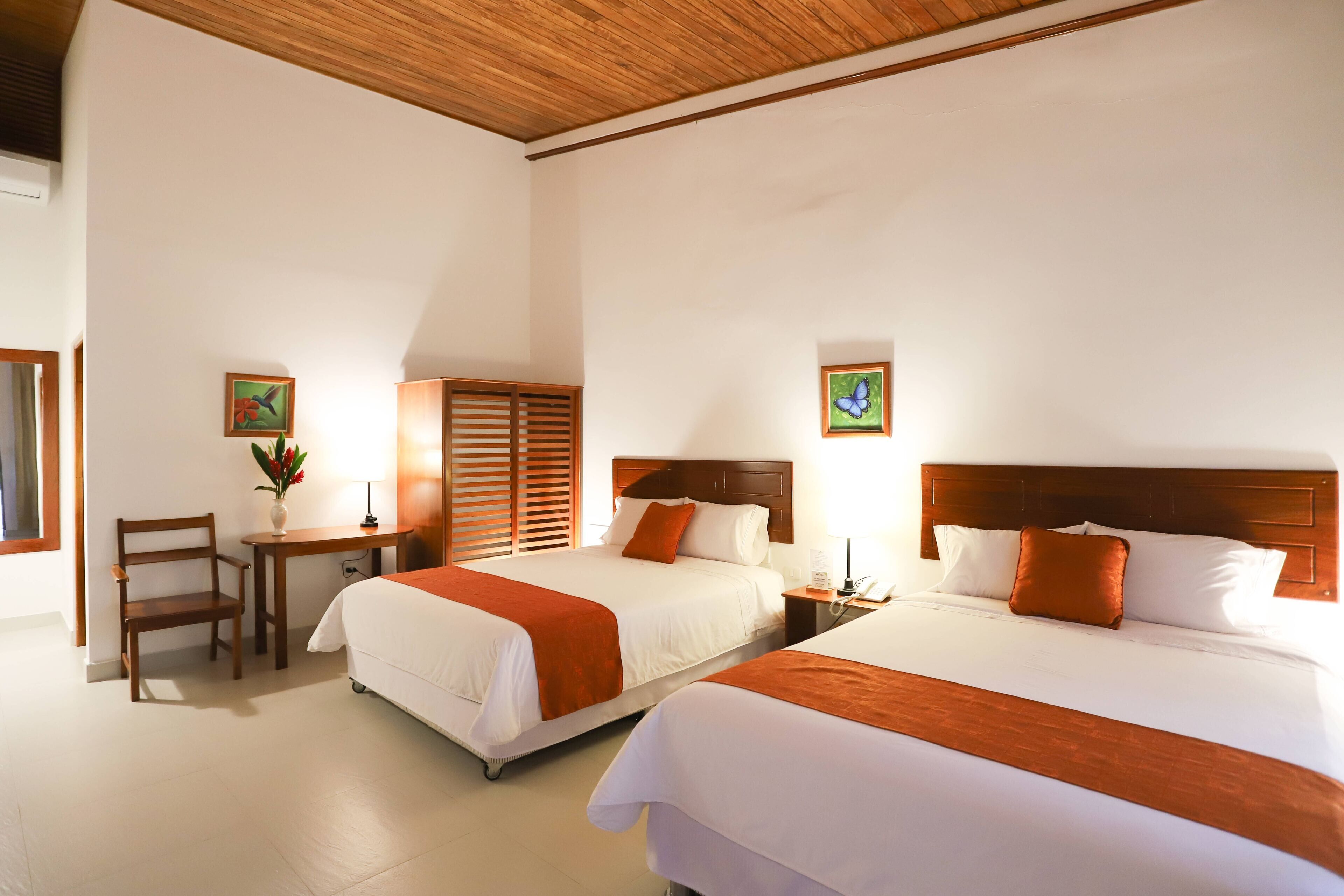 Une chambre d’hôtel avec deux lits, des têtes de lit en bois, un bureau avec une chaise et une petite table sur laquelle est posé un vase de fleurs.