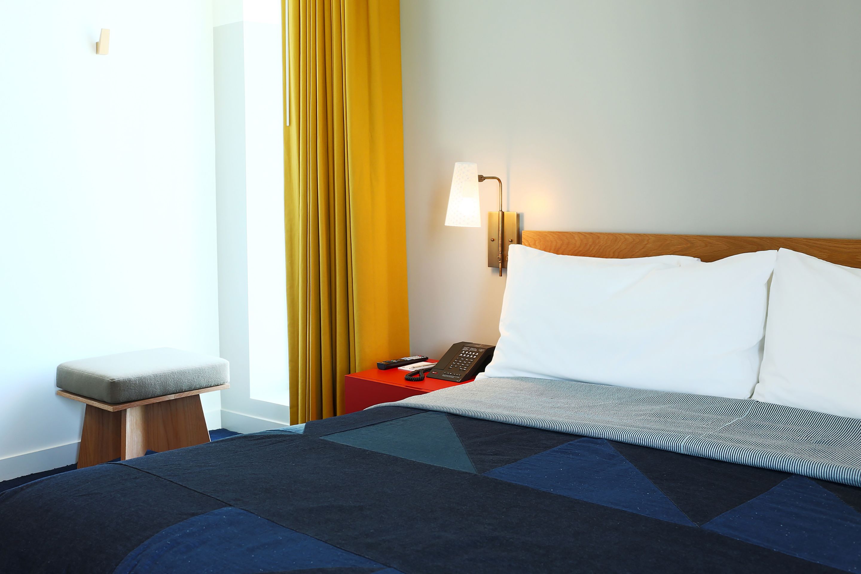A hotel room with a bed, a bedside table with a phone, a lamp, and a small stool.