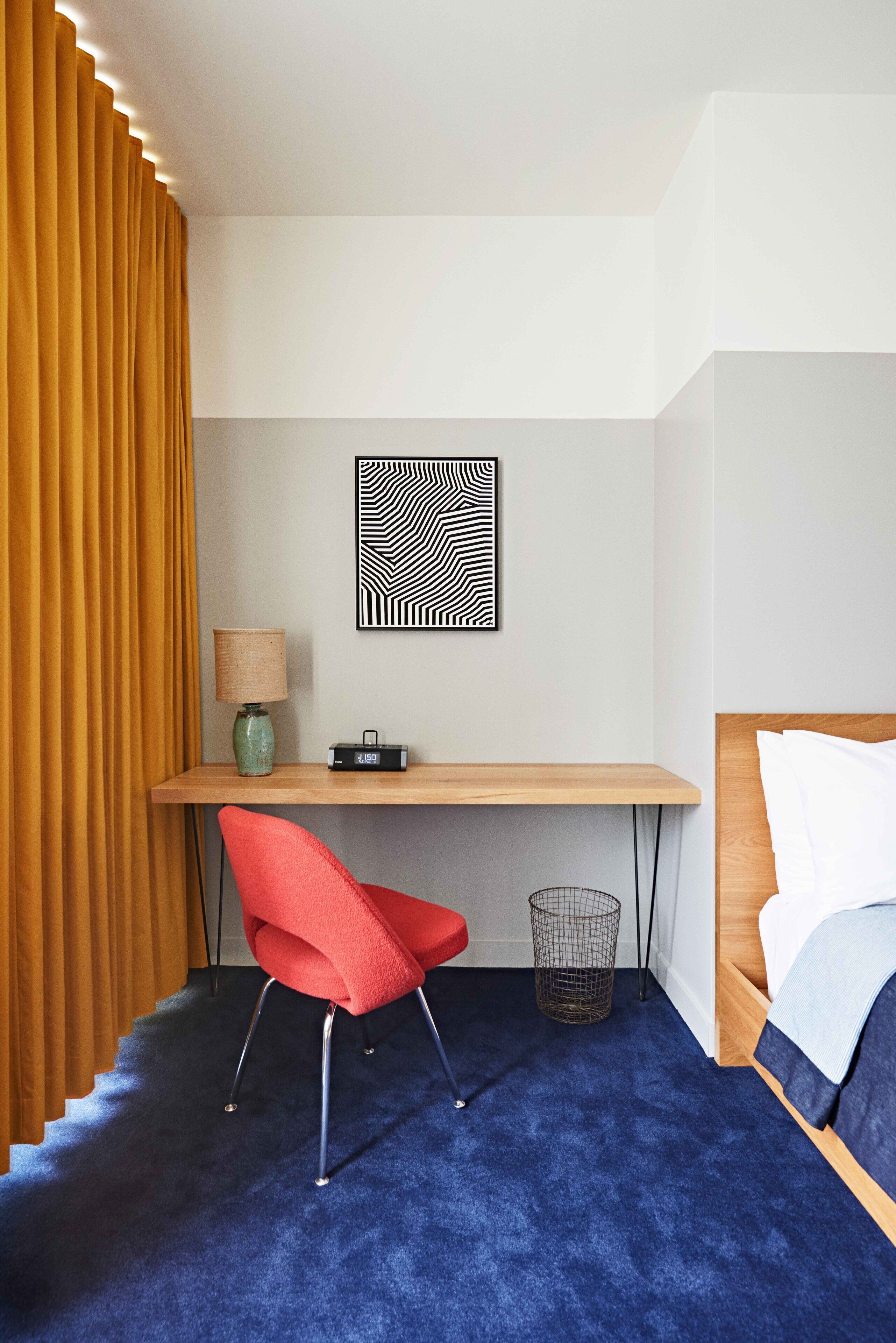 A hotel room with a wooden desk, a red chair, a lamp, a radio, a framed abstract art piece, a bed with white bedding, and a blue carpet.