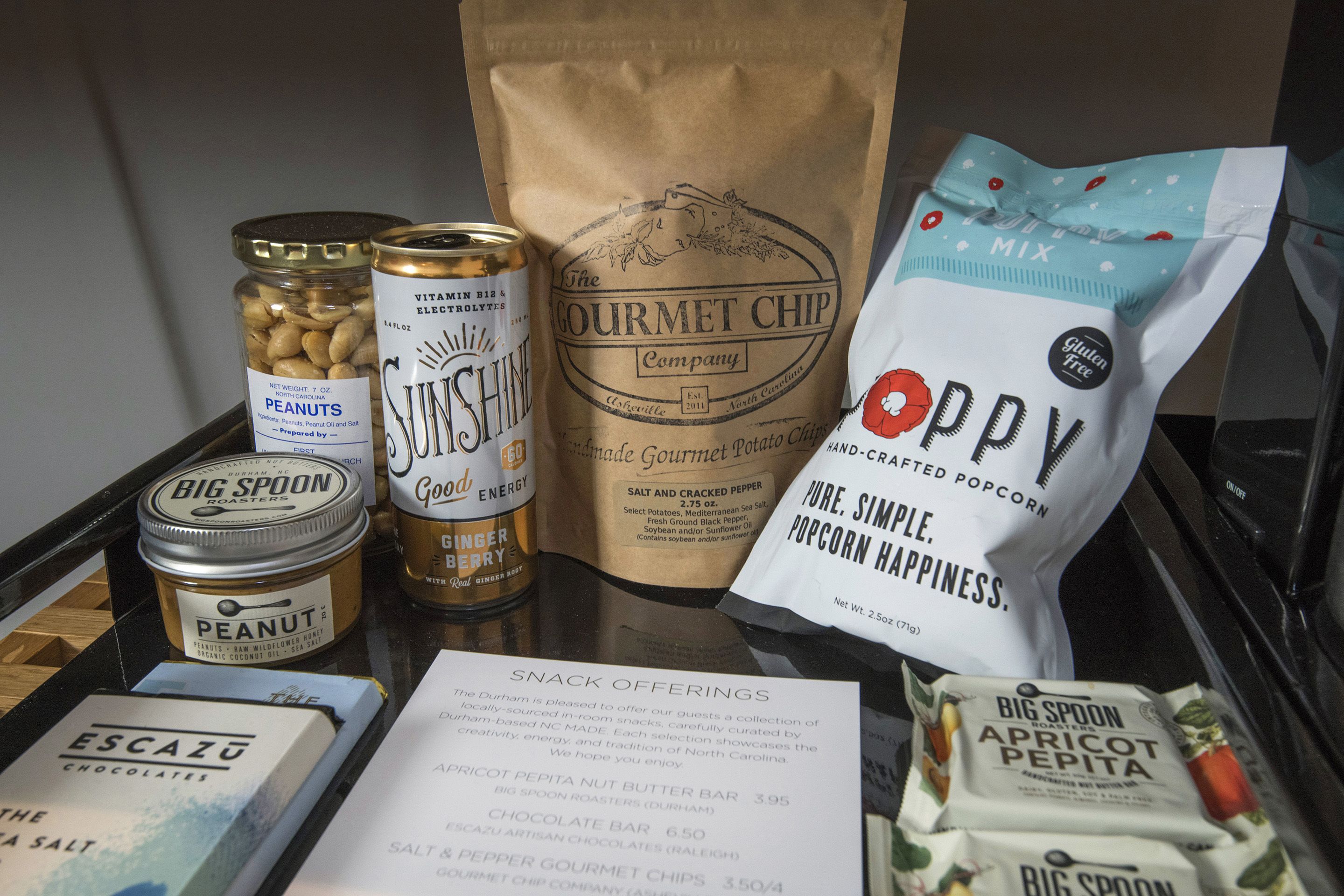 Eine Serviertablett mit verschiedenen Leckereien, darunter eine Erdnussglase, eine Energydrinkdose, eine Tüte Gourmet Chips, eine Tüte Mohn-Popcorn und eine Schachtel Escazu Pralinen.