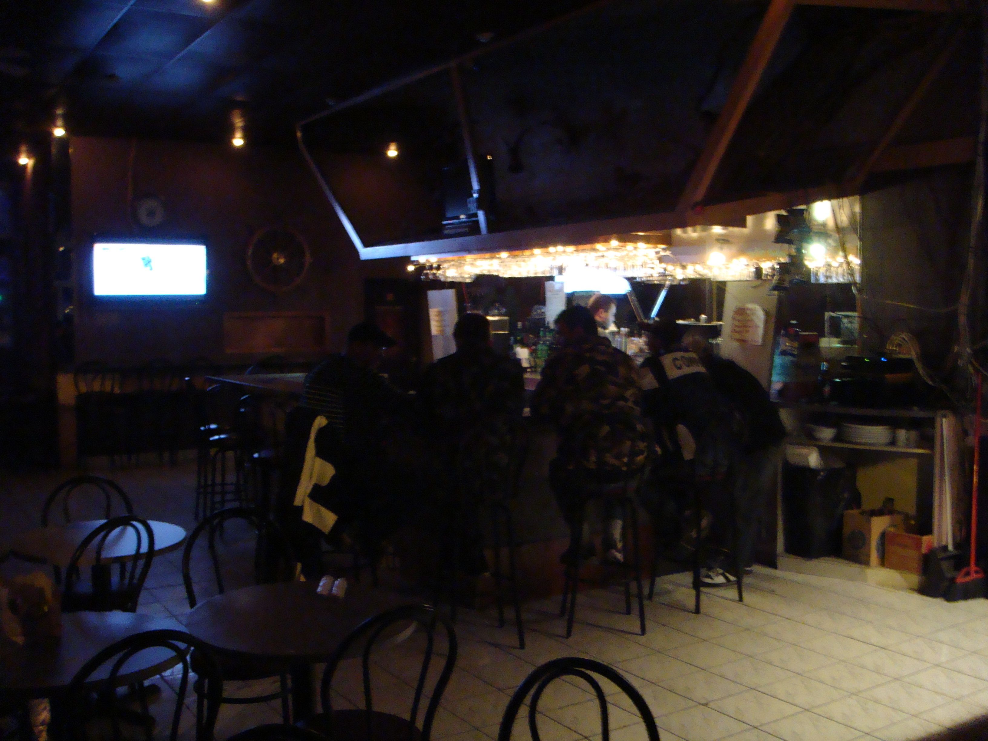 Uma área de bar com pouca luz, mesas, cadeiras, um balcão com um bartender e uma tela de televisão.
