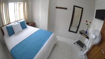 A hotel room with a bed, a bedside table, a mirror, a TV, and a fan.