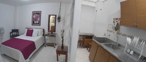 A compact hotel room with a bed, bedside table, and a small desk with a chair. There is a kitchenette with a sink and a dish rack.