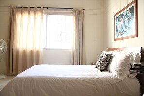 Ein Hotelzimmer mit Bett, Nachttisch, Ventilator und einem fenster mit Vorhängen.