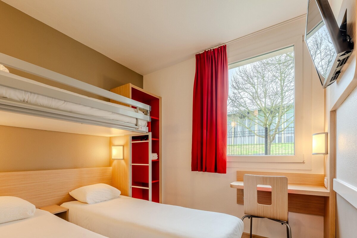 A hotel room with two beds, a desk, a chair, a TV, and a window with a view of trees.