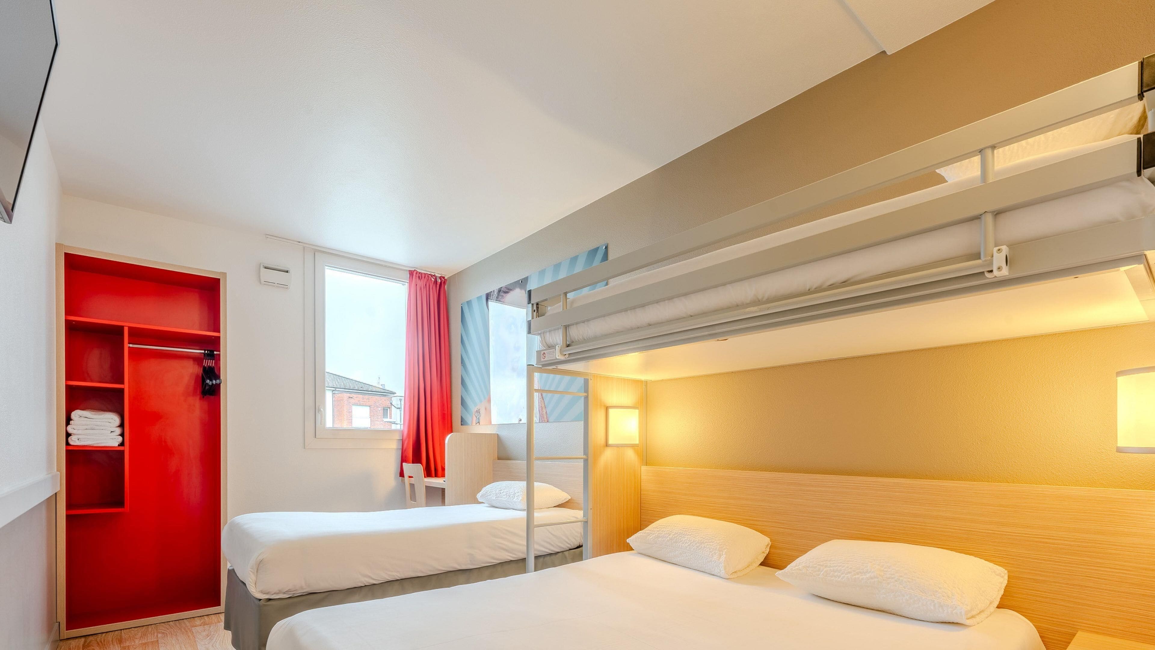 Hotellihuone, jossa on kerrossängyt, punainen vaatekaappi, ikkuna, josta on näkymä rakennukseen, ja yöpöytälamppu.