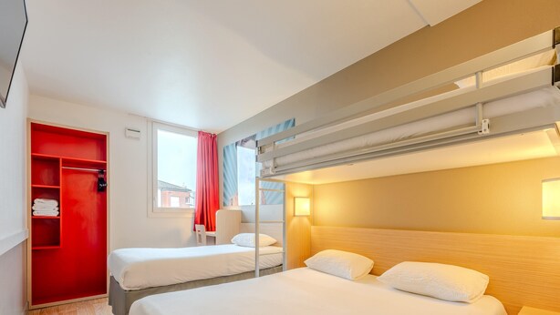 Habitación de hotel con literas, un armario rojo, una ventana con vista a un edificio y una lámpara de noche.