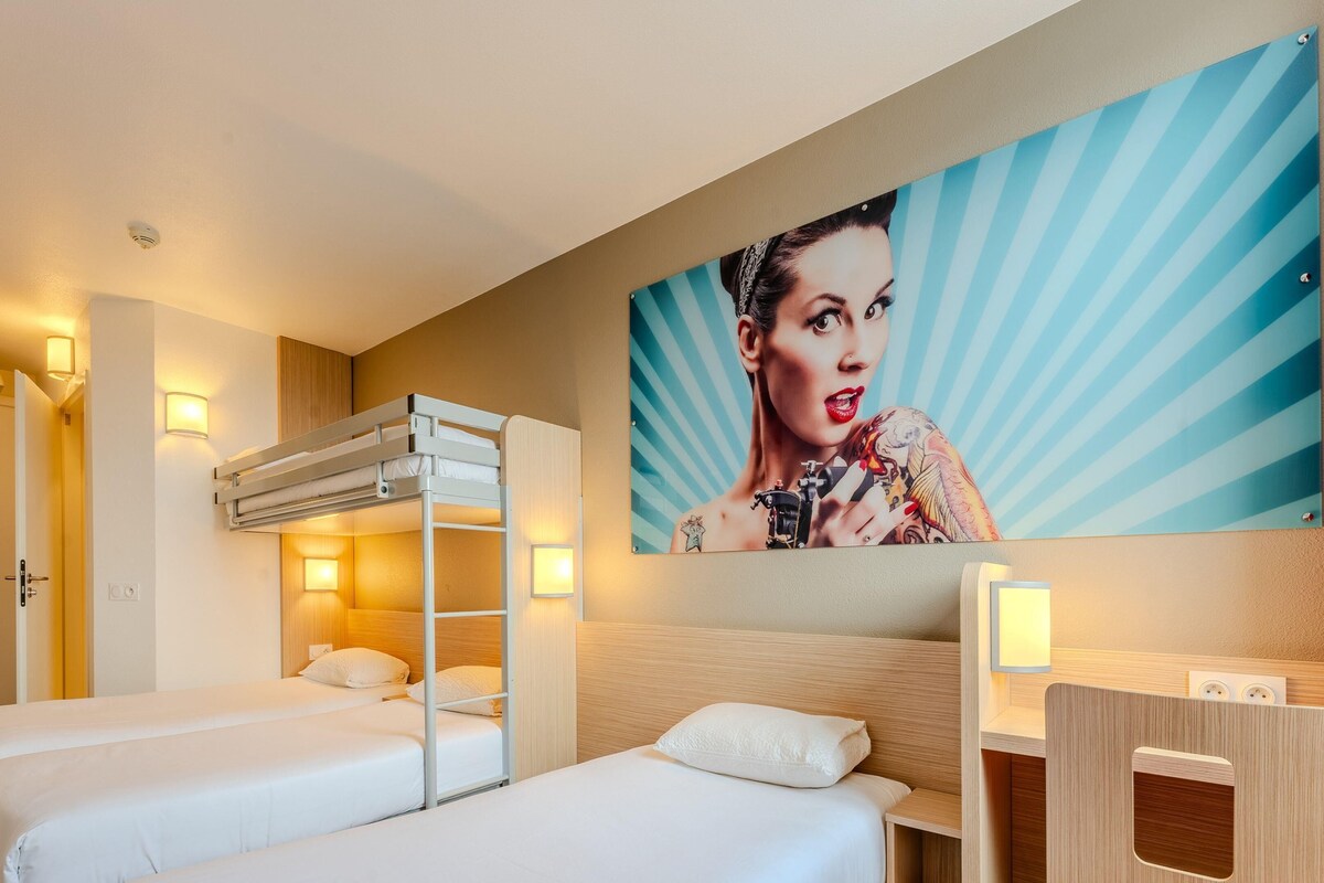 A hotel room with two beds, a bunk bed, a large wall mural, a desk with a chair, and a wall-mounted air conditioner.