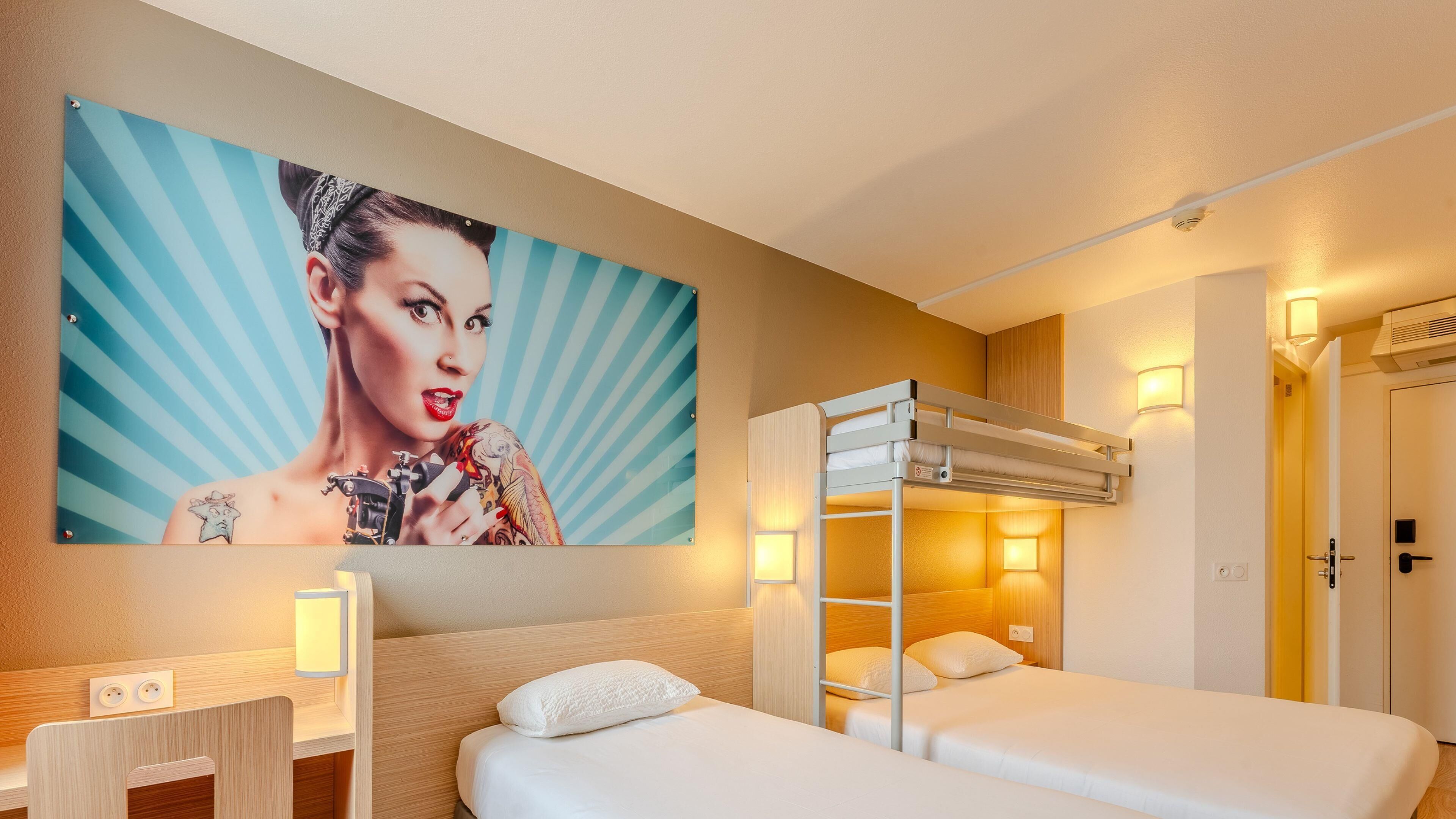 A hotel room with a bunk bed, a bedside table, a lamp, a painting on the wall, and a door.