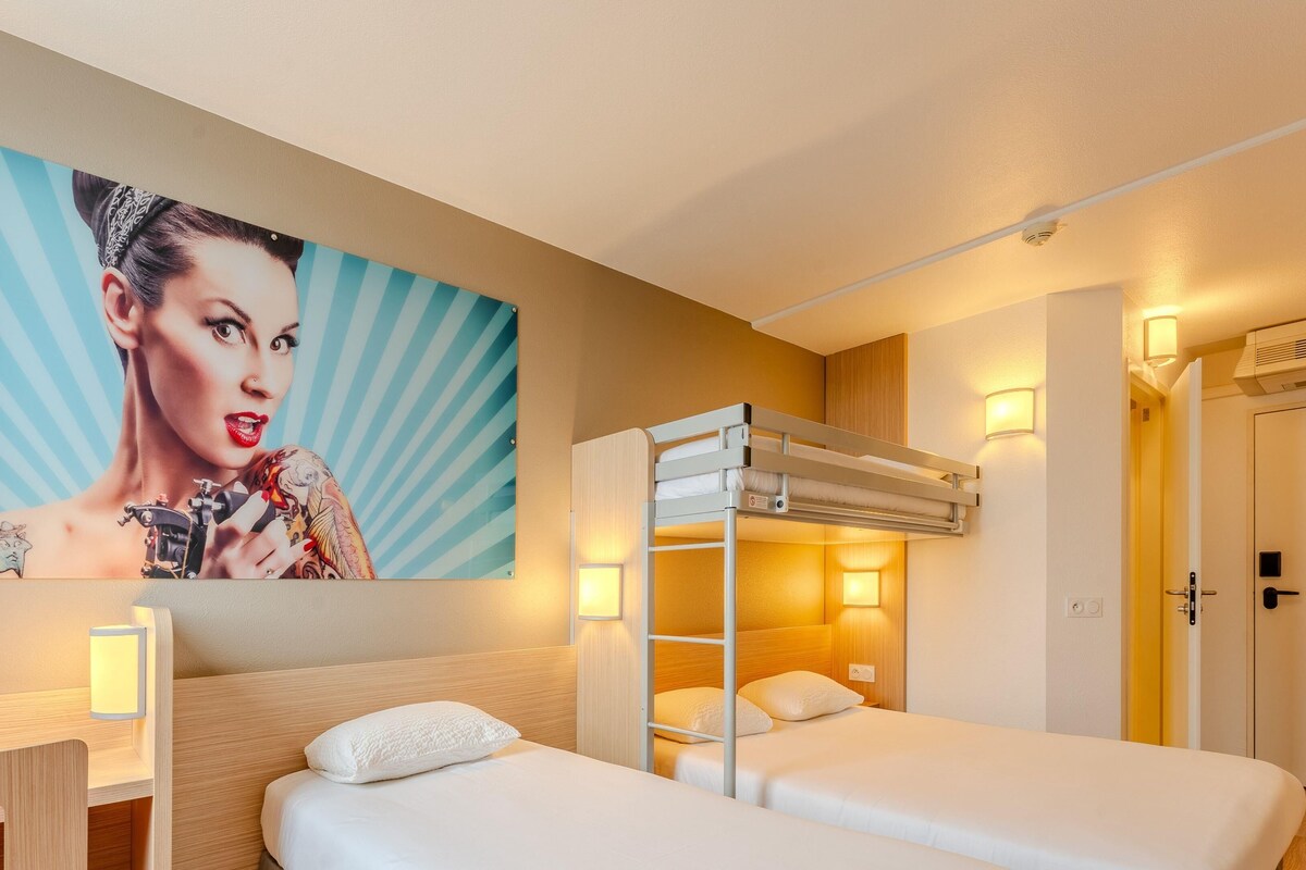 A hotel room with a bunk bed, a bedside table, a lamp, a painting on the wall, and a door.