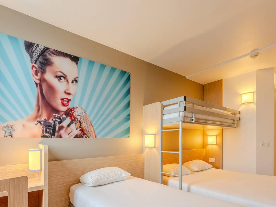 A hotel room with a bunk bed, a bedside table, a lamp, a painting on the wall, and a door.