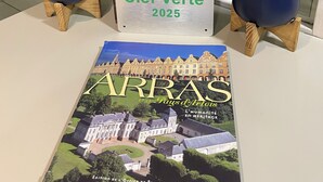 Un folleto titulado 'ARRAS' con una ilustración de un edificio, un letrero con forma de llave que dice 'Clef Verte 2025' y dos plantas en macetas sobre una mesa.