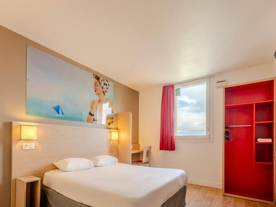 Habitación de hotel con cama, escritorio, silla, televisor, armario rojo y ventana con vista al cielo.