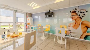 Un comedor amplio e iluminado con mesas y sillas blancas, un mural en la pared y vistas al exterior a través de amplias ventanas.