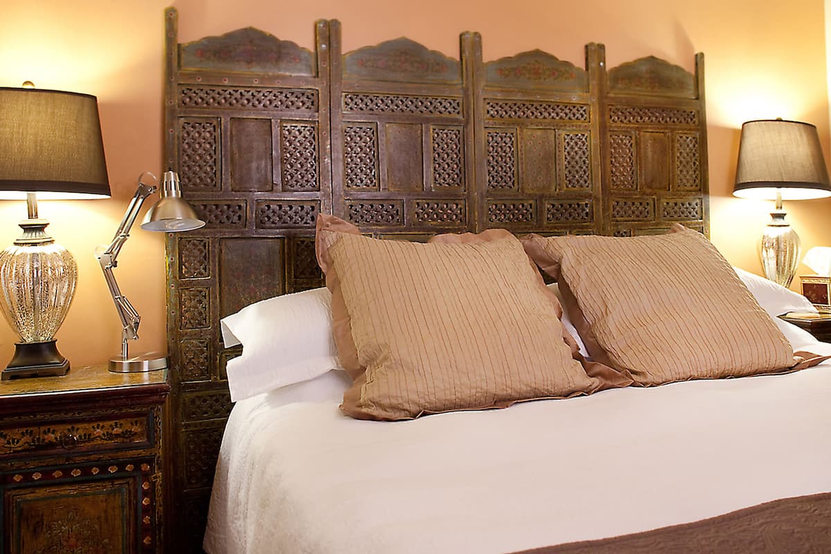 Cama bem arrumada com duas almofadas, cabeceira de madeira com entalhes elaborados e duas luminárias de cabeceira de cada lado.