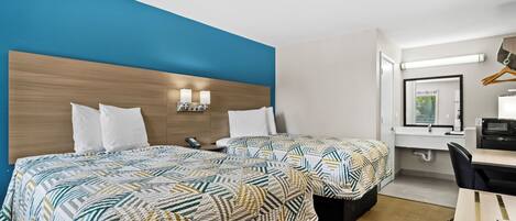 Quarto de hotel com duas camas, uma escrivaninha, uma cadeira, um espelho e um banheiro.