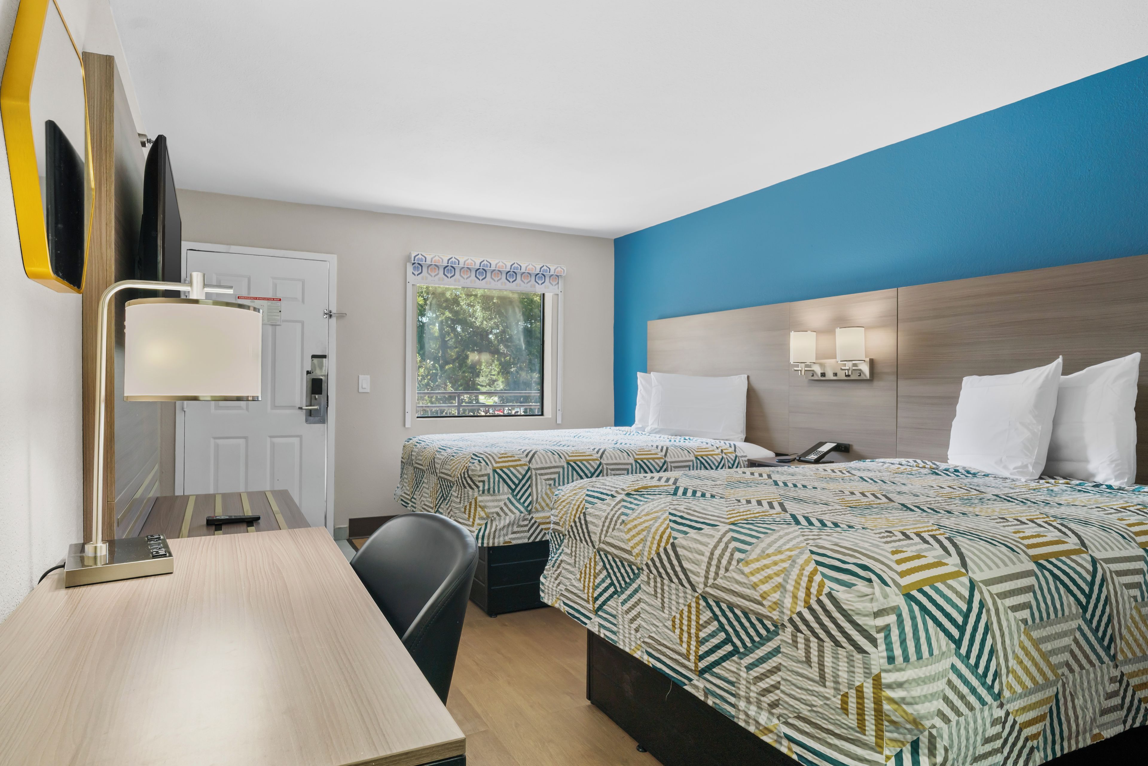 A hotel room with two beds, a desk, a chair, a desk lamp, a window with a view of trees, and a door with a sign.