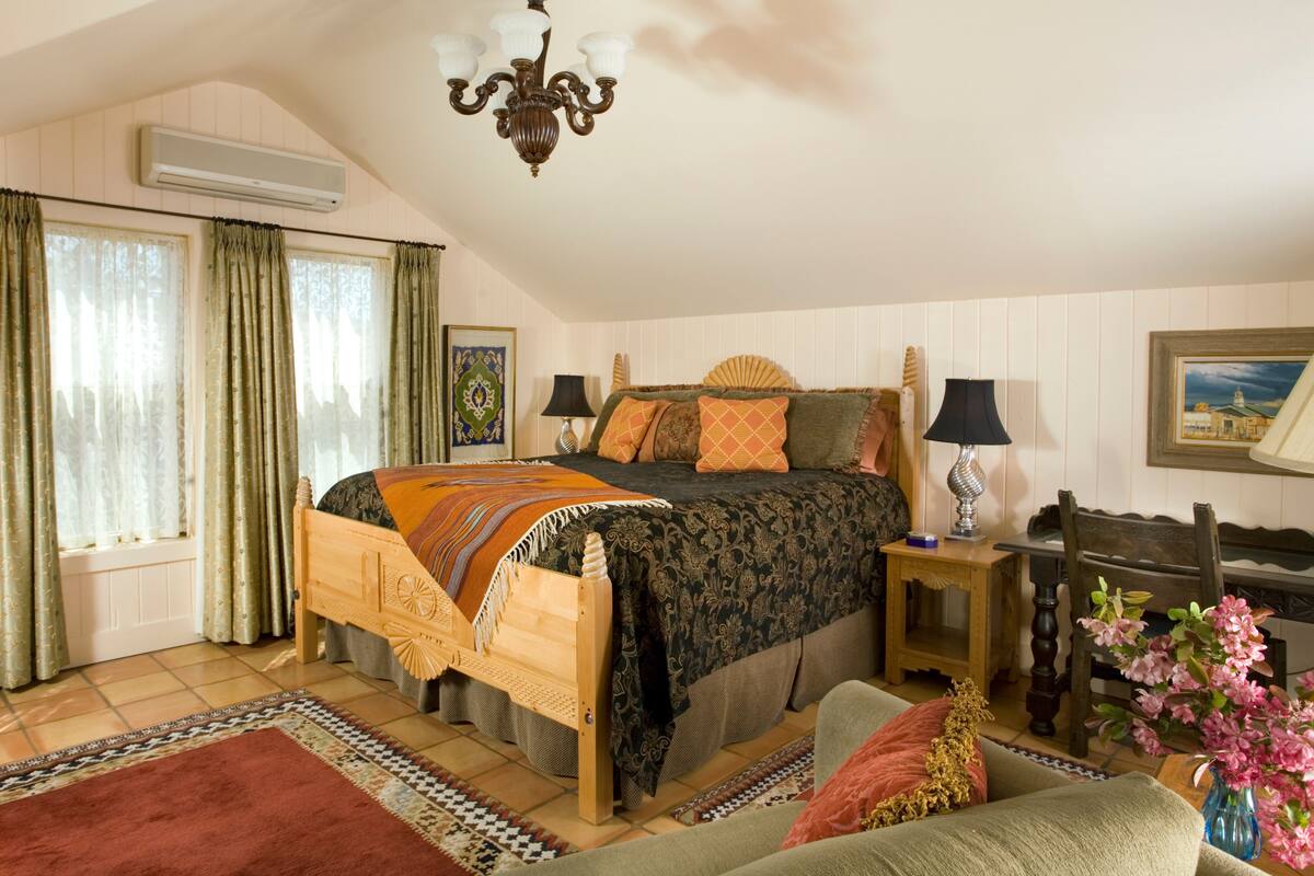 A bedroom with a wooden bed, two bedside tables, a chandelier, a window with curtains, a framed picture on the wall, and a floral arrangement on the floor.