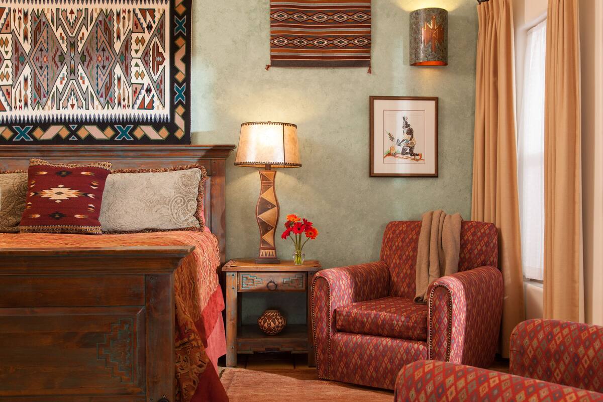 A traditional room with a bed, a patterned armchair, a side table with a lamp, and a wall hanging.