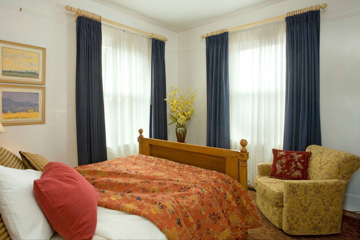 A hotel room with a bed, a chair, a lamp, and two windows with curtains.