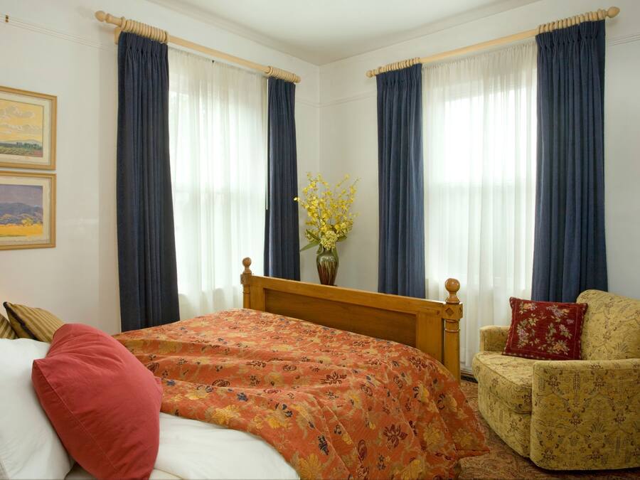 Una habitación de hotel con una cama, una silla, una lámpara y dos ventanas con cortinas.