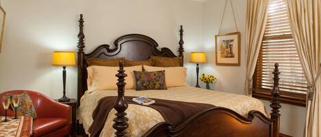 A bedroom with a four-poster bed, two bedside lamps, a red chair, a small table with a lamp, and a window with curtains.
