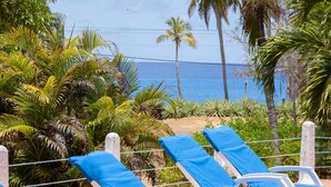 Fileira de cadeiras de praia brancas com almofadas azuis, uma mesa com bebidas e vista para o oceano e palmeiras.